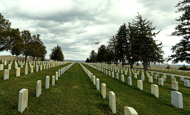 Photo of the Custer National Cemetery looking south.  Rows of white headstones are seen in the green grass.