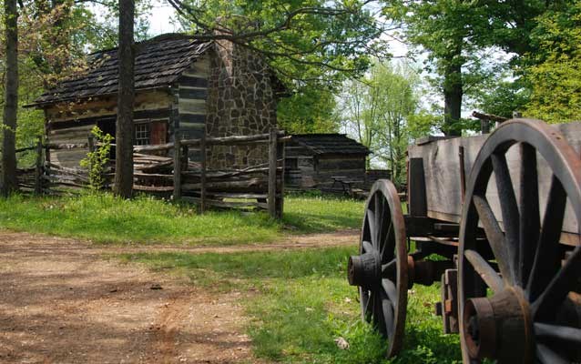 wooden wagon just off right of tree lined trail leading to log cabin.