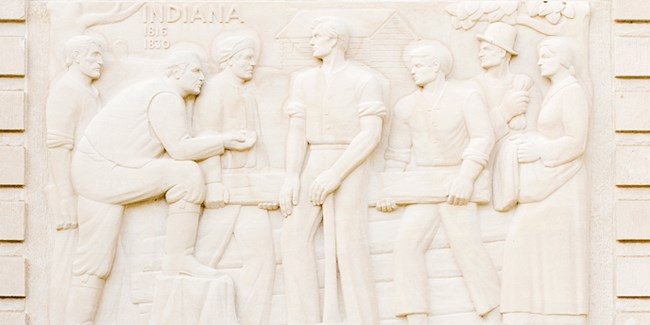 Scene carved in limestone showing seven people, three on each side of young man holding an axe. Text, Indiana 1816-1830.