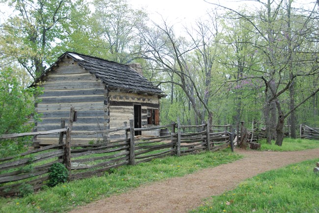 Rock trail leads to log cabin. Split rail fence between trail and cabin. Trees in background.