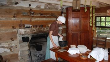 Girl in pioneer dress in log cabin putting food on plate at wooden table. Hearth and cabinet behind her.