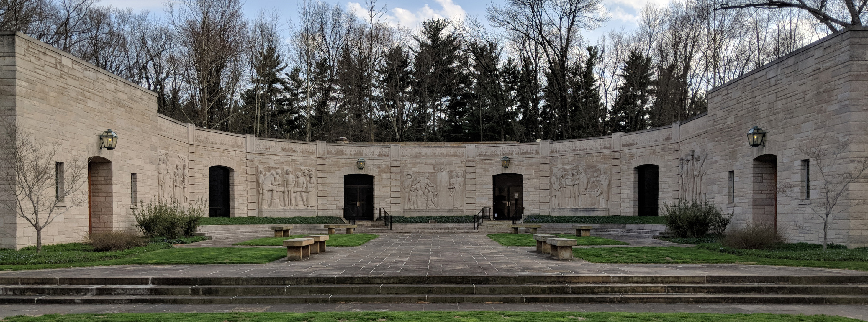 Curved limestone building with 5 sculptured panels with scenes of people, stone walkway leads to stairs and doorway, grass between walkways, stone benches