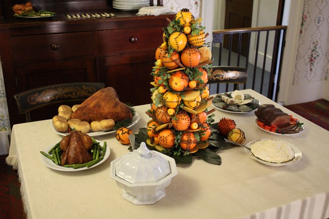 Dining table with ham and rolls; chicken and asparagus; mashed potatoes, roast beef, oysters; and a tower of oranges adorned with leaves, cloves, and cinnamon sticks