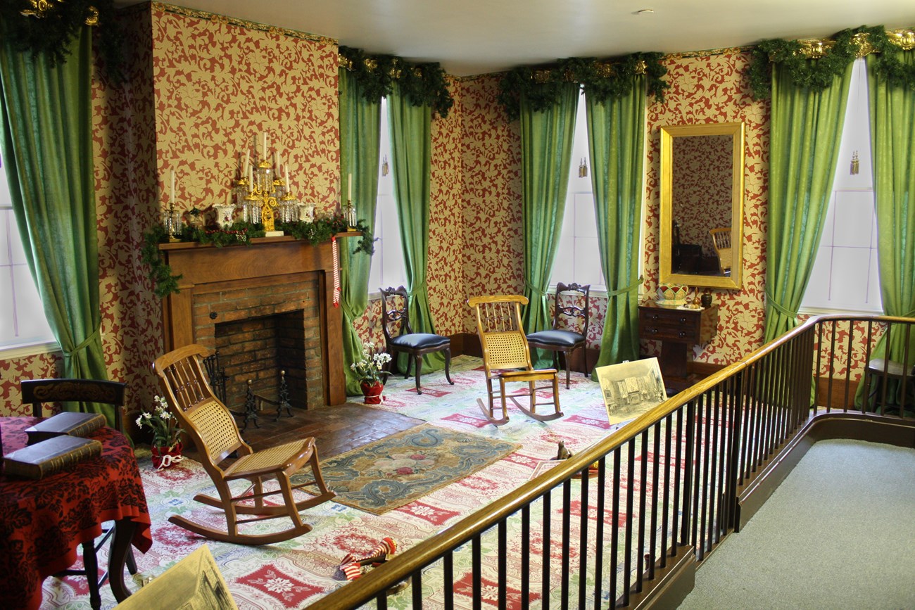 Lincoln Home sitting room decorated for Christmas with evergreen draped over fireplace mantle and golden window valences. A striped red and white stocking hangs from mantle. Small toys are scattered on the floor.