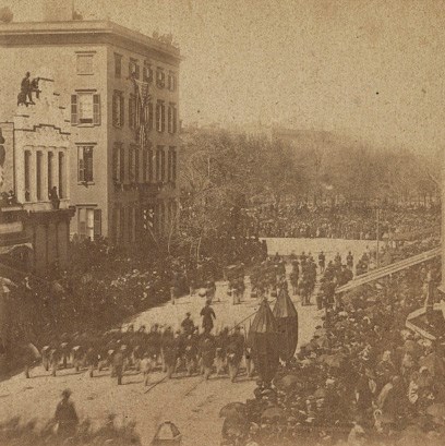 View of funeral parade in the street from higher building. Everyone is wearing black. A 4-story boxy building and 2 story building with pillars is seen across the street on the left, with people watching from the windows.