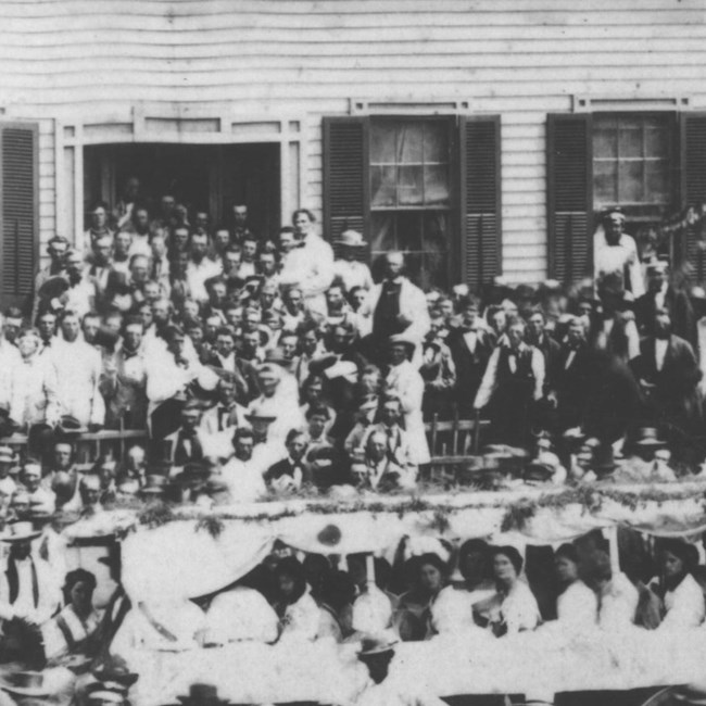 Close up of 1860 rally photo of Abraham Lincoln, wearing white, on steps of his house surrounded by admirers. He towers over everyone around him.