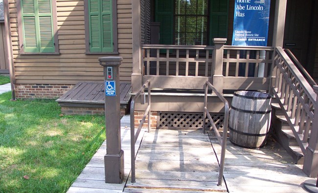 Wheelchair lift next to outdoors steps that lead to a porch. Lift has safety railings on left and right sides. On left of lift is wooden rod with buttons for controlling the lift.