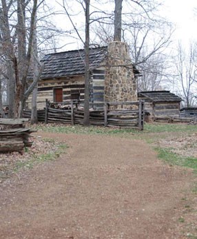 cabin with stone chimney at end of brown driveway that curves left with split rail fence