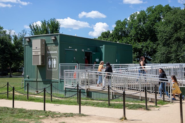 Visitors walk up a metal ramp to a green rectangular restroom building.