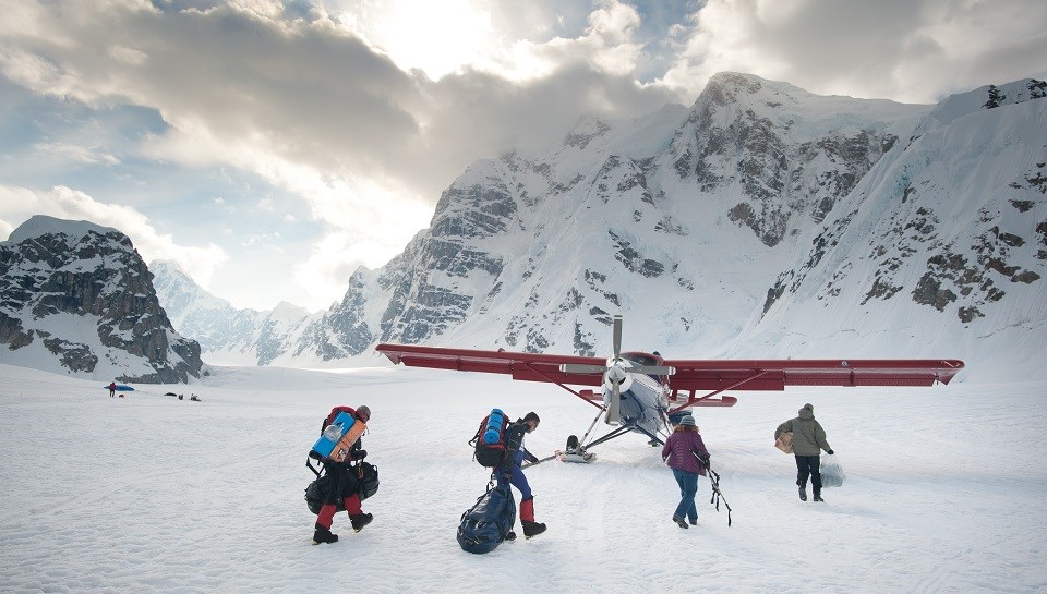 Four hikers on a snowy mountain walk with gear to a plane on skis.