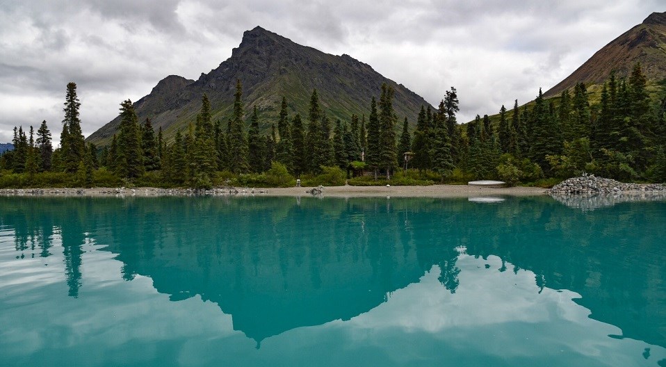 Green and brown mountains, spruce trees, a wooden cabin and a metal canoe reflect in a turquoise lake.