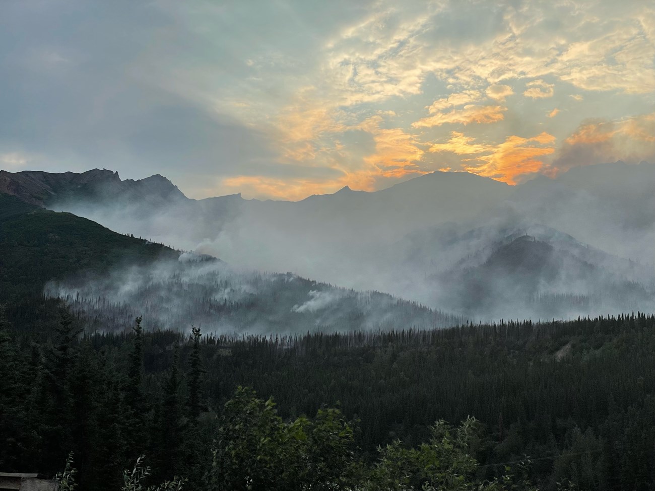 Smoke rises off of forested mountains while the sun sets in the sky