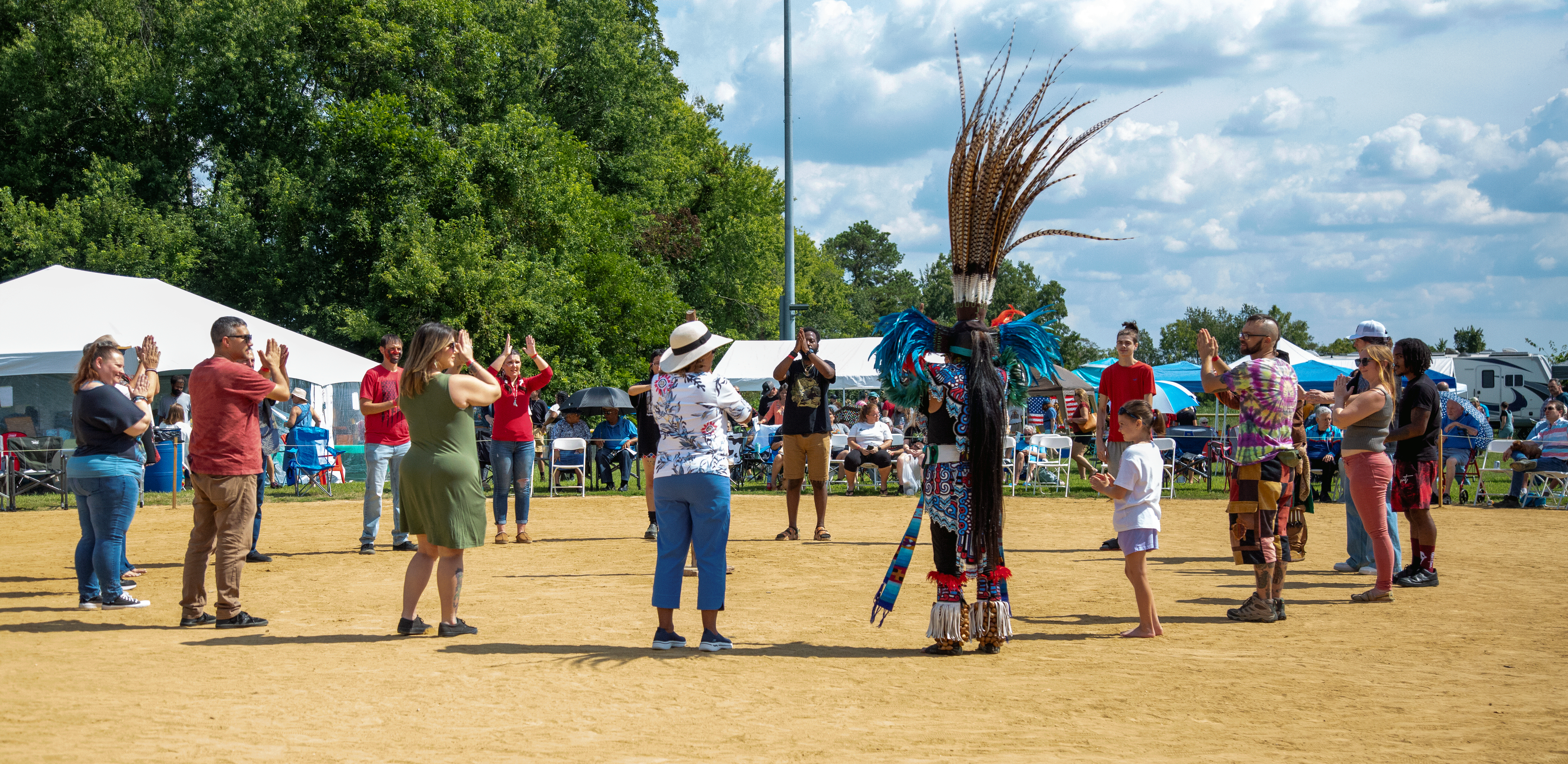 Group dancing around an individual in traditional attire with elaborate headdress at an outdoor event.