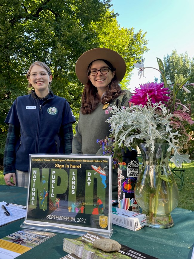 Intern and park ranger stand behind a table with flowers and a sign reading "National Public Lands Day"