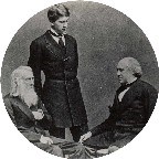 historic black and white photo of three men. Two older men sitting down facing one another and a younger man standing between them.