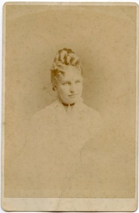 Bust-length portrait of a young woman. Her hair is worn in an elaborate coiffure of coils and a comb.  A dark ribbon with hanging pendant is around her neck.  Her outfit is very-light colored.