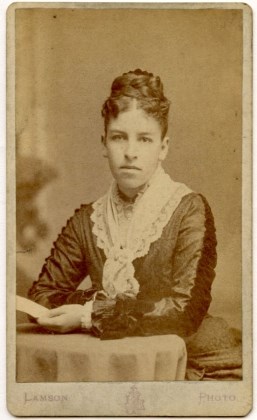 Half-length portrait of a young woman sits at a table holding a letter in her left hand.  She wears a dark-colored dress with lace kerchief (a black ribbon is around neck.)  A braid is coiled around her head.