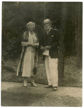 An older man and woman stand on path in front of evergreen trees.  He holds papers in his hands.
