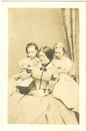 A young woman in 19th-centry dress reads a book while two girls look over her shoulders from the left and right.