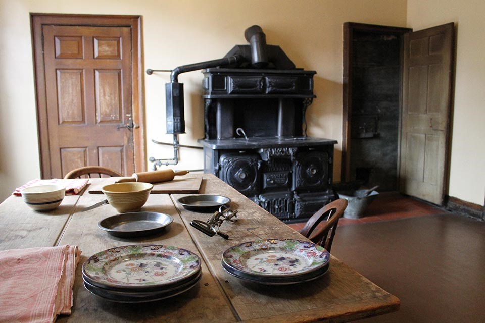 View of kitchen with large coal stove in background, table with plates and kitchen tools in foreground