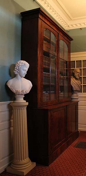 Floor to ceiling bookcase flanked by a marble bust of angel with flowers and plaster bust of Henry Longfellow