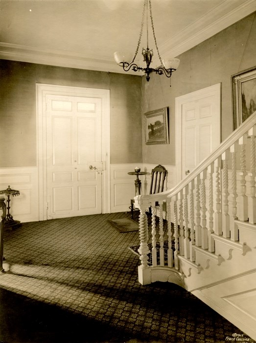 Black and white photograph of a hallway with stairs in the foreground and a large door on the opposite wall.
