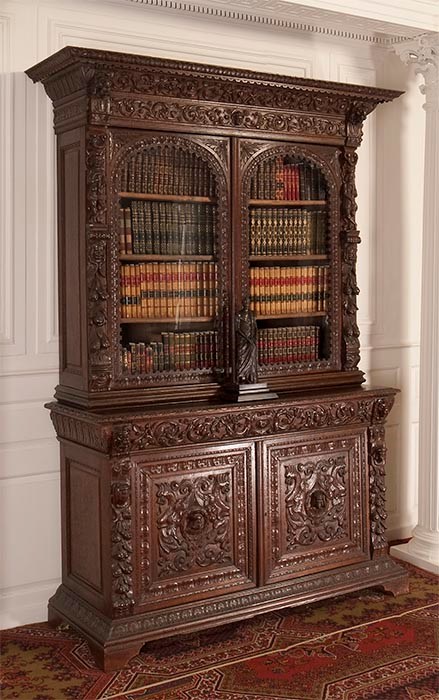 Tall, ornately carved bookcase. Upper case with arched glass windows showing four shelves of books. Lower case with ornately carved wooden doors.