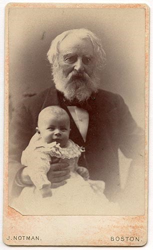 Bearded man in suit seated, holding baby in long white dress on his lap