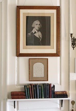 Framed print of portrait of Washington hanging above framed manuscript page and shelf of books
