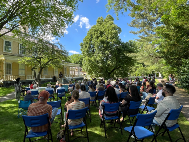 Poet reading on stage with audience outside on the lawn.