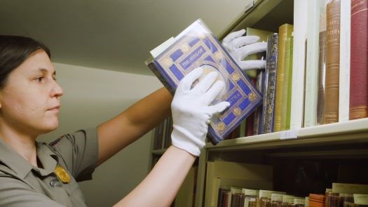 Woman wearing white gloves and a National Park Service uniform places a historic copy of "The Song of Hiawatha" on a bookshelf