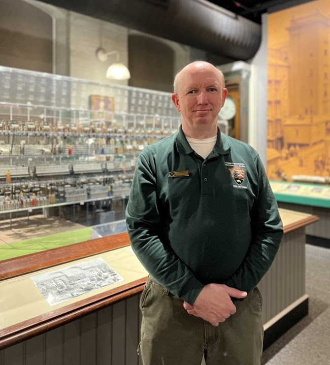A man wearing a long sleeve, green volunteer shirt stands in front of a mill model in a museum space.
