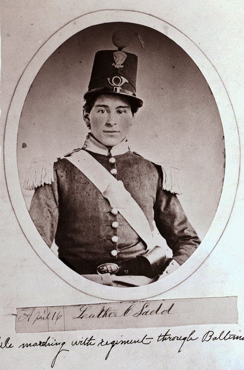 A black and white portrait of a young man wearing an intricate unifrom.  Below the words read 'April 16, Luther C Ladd, Marching with regiment through Baltimore'.