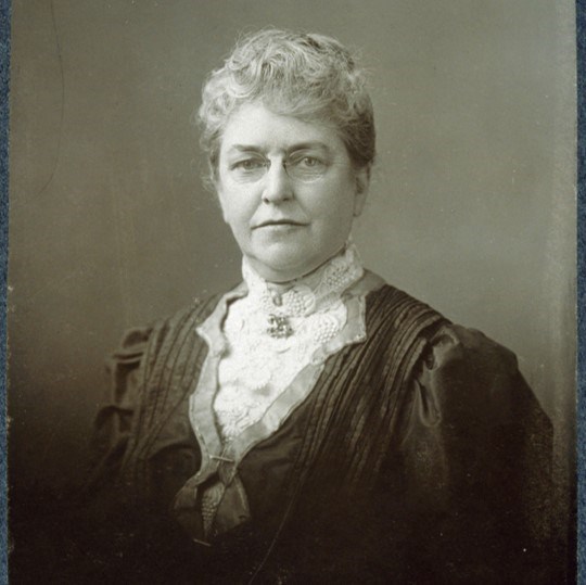 A grayscale portrait of an older woman dressed in late 19th century dress looking straight out.