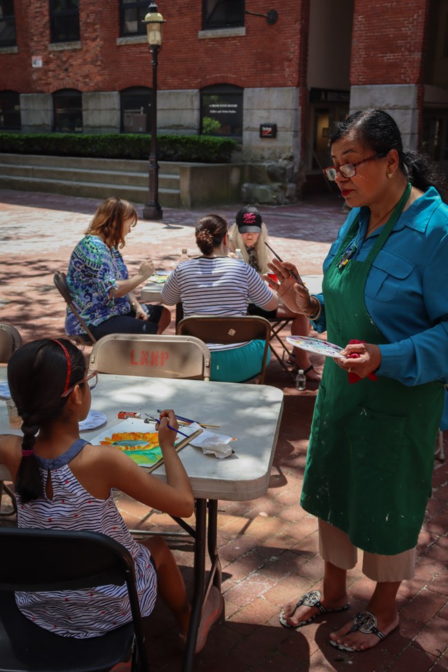 An artist instructs a young student as visitors sit around tables painting with watercolor.