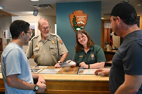 Two people, one in park service uniform, the other in a green shirt with arrowhead patch, talk to people from behind a desk.