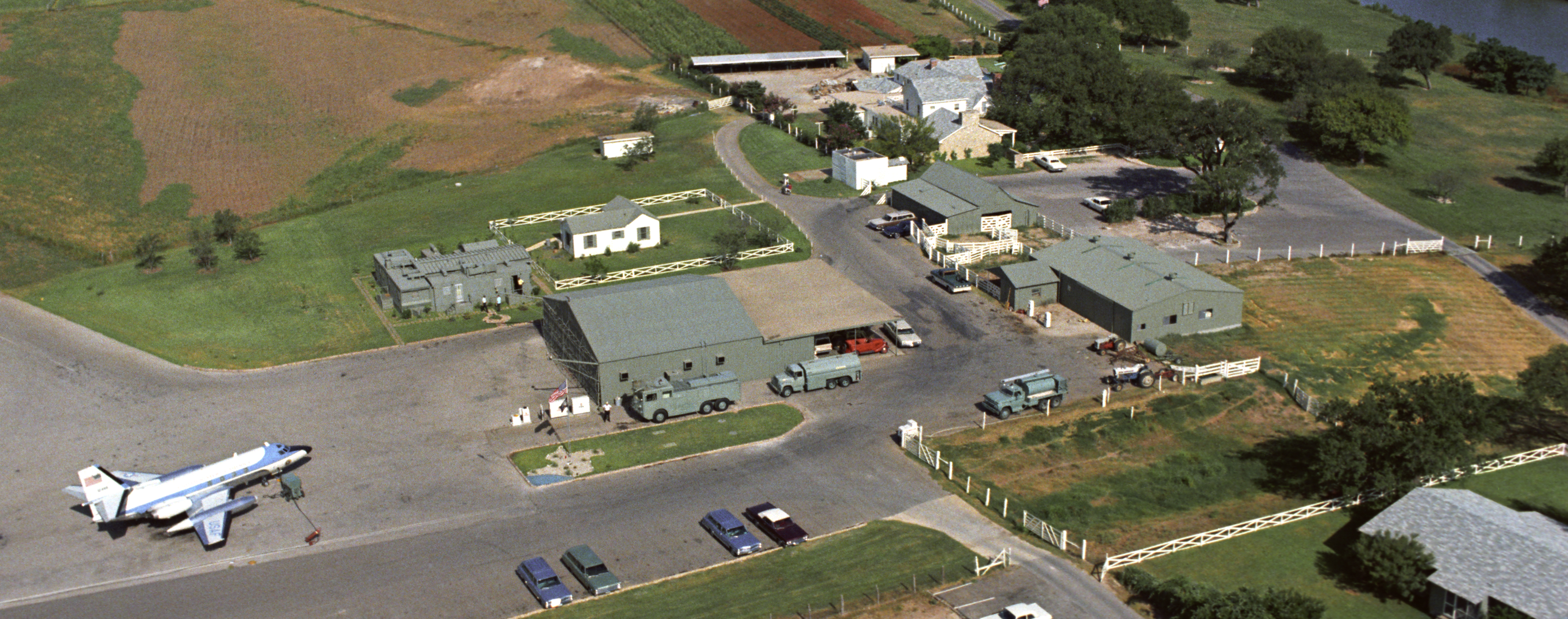 AN aerial view of numerous green buildings and a jet, surrounded by grassy fields.