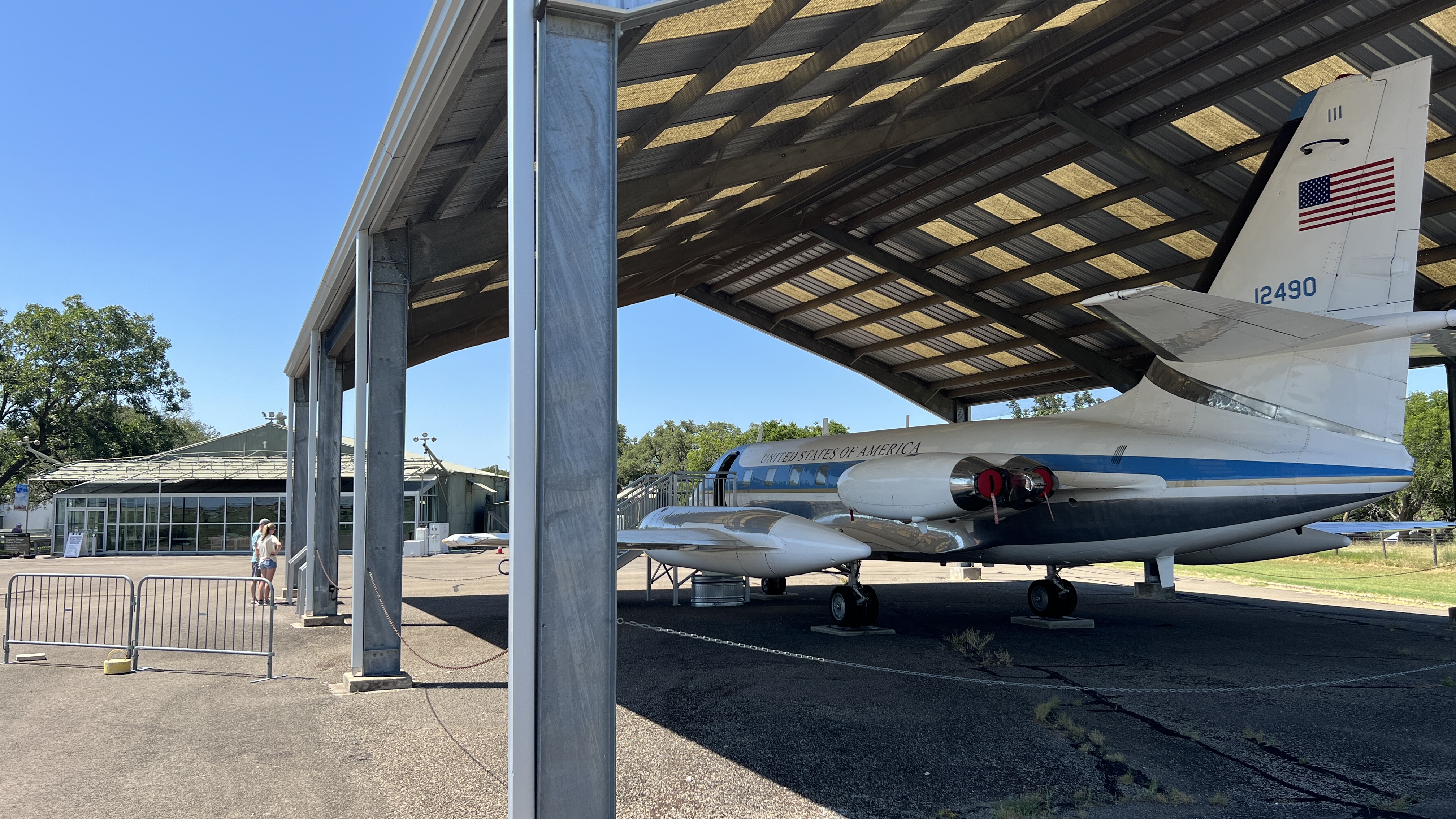 Two people are reading a sign while standing underneath a large open shed housing a jet airplane.