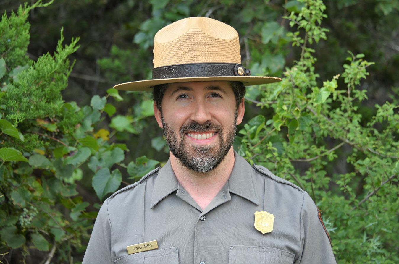 Bearded ranger in uniform with badge and straw flathat. Background of leafy green bushes.