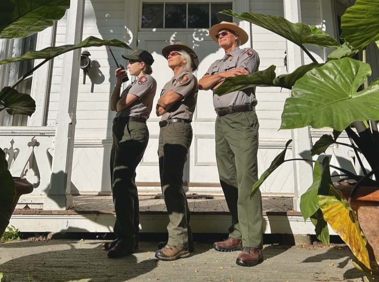 three park rangers stand crossing arms facing left side of image in front of large potted plants and ornate white building with columns