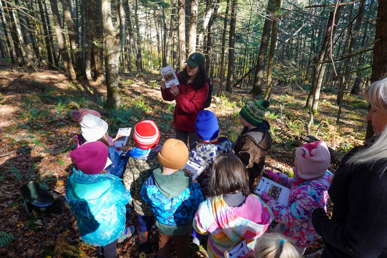 instructor in ball cap talks to preschool aged children dressed warmly on a trail