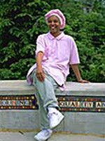 A smiling woman in a pink shirt and pink head covering sits on a concrete wall, decorated by colorful art.