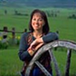 A dark-haired woman in a blue shirt leans against an old wooden wagon wheel, with green pastures behind her.