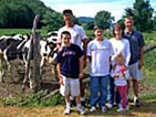 A family of six stands in front of a wooden fence, with black and white Holstein cows grazing behind them.