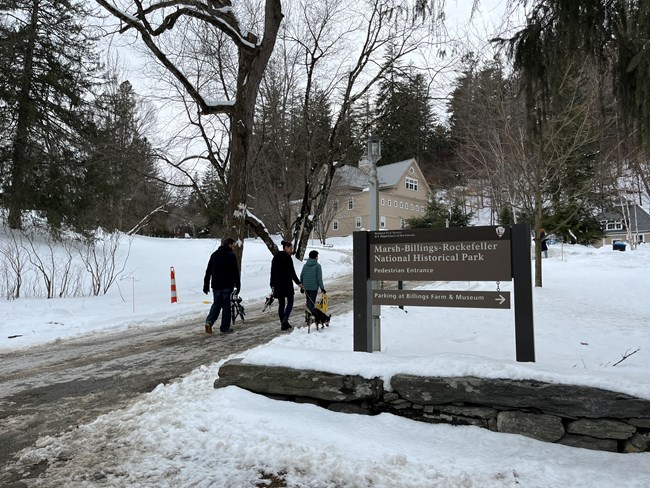 family of three walks up trail holding snowshoes next to marsh-billings-rockefeller sign