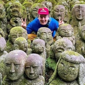 man with red hat and blue shirt holds up peace signs among moss covered statues with both hands