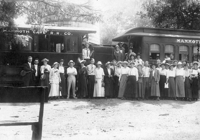 A black and white photo of a large group of people wearing early twentieth century clothing standing in front of two train cars with painted text “Mammoth Cave R. R. CO.” along their sides.
