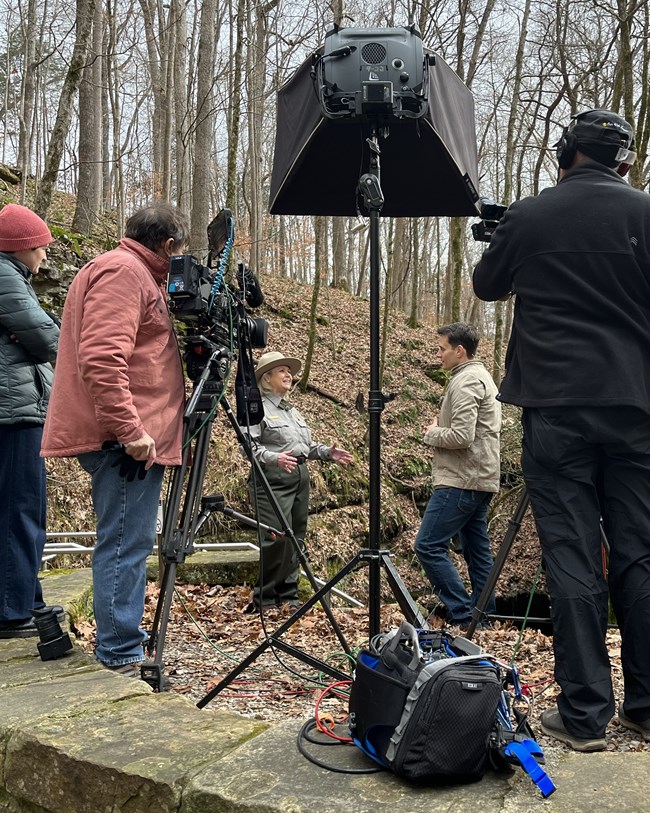 A film crew with movie cameras and large lights film two people talking in the woods.
