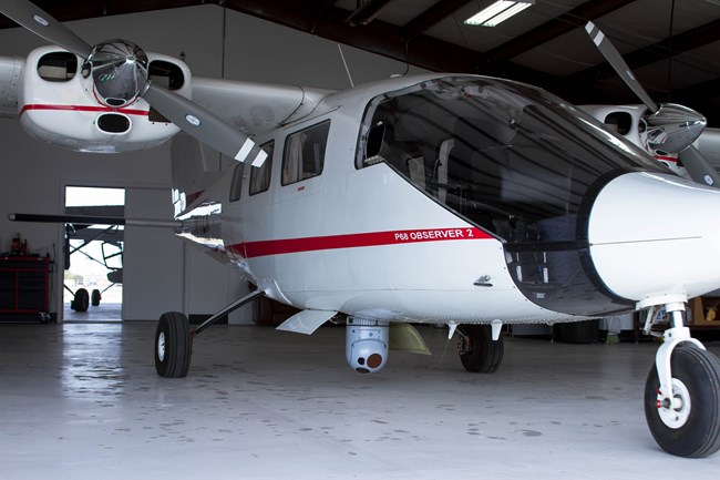 A small, two-engine airplane with camera equipment attached to it is parked inside an airplane hangar.