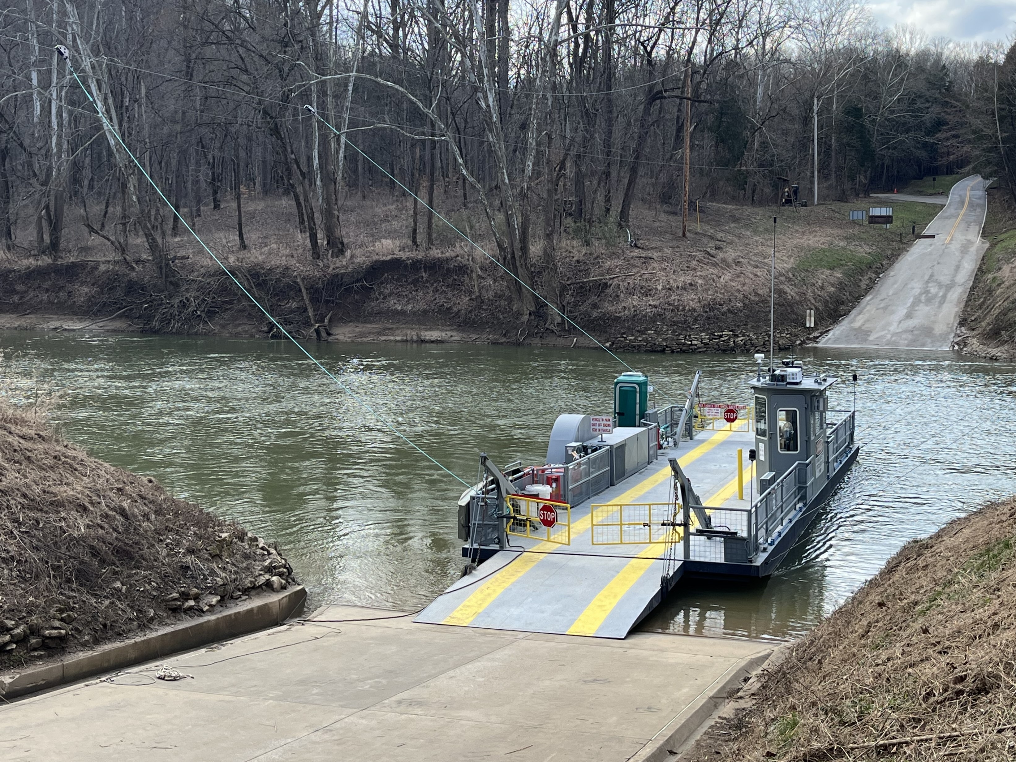 A vehicle ferry travels across a river toward a roadway with a car approaching the water’s edge.